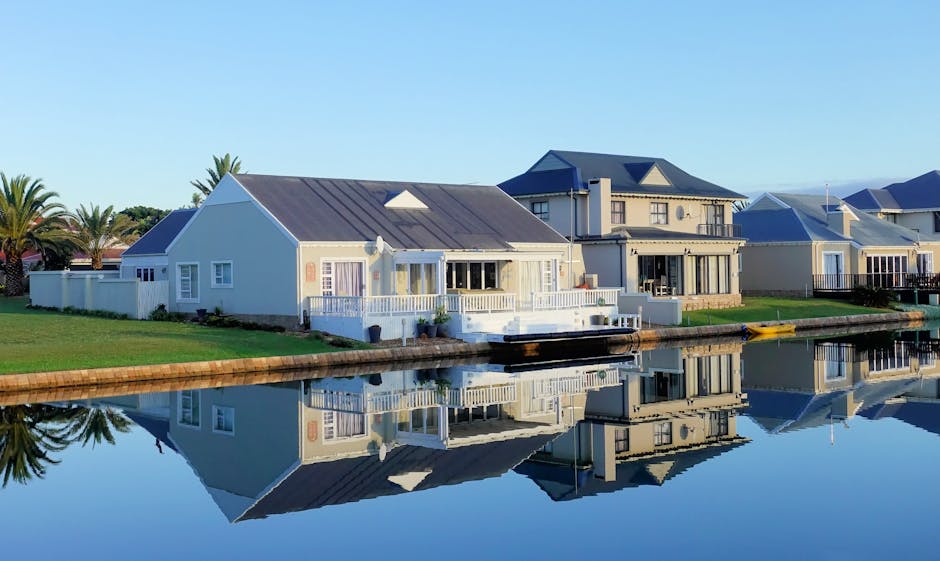 Luxury modern homes reflecting on a peaceful, clear lake during daylight, showcasing architectural beauty